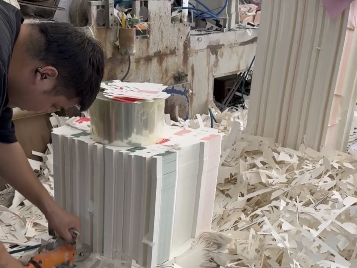 Factory worker trimming white molded packaging pieces beside stacked materials and paper offcuts